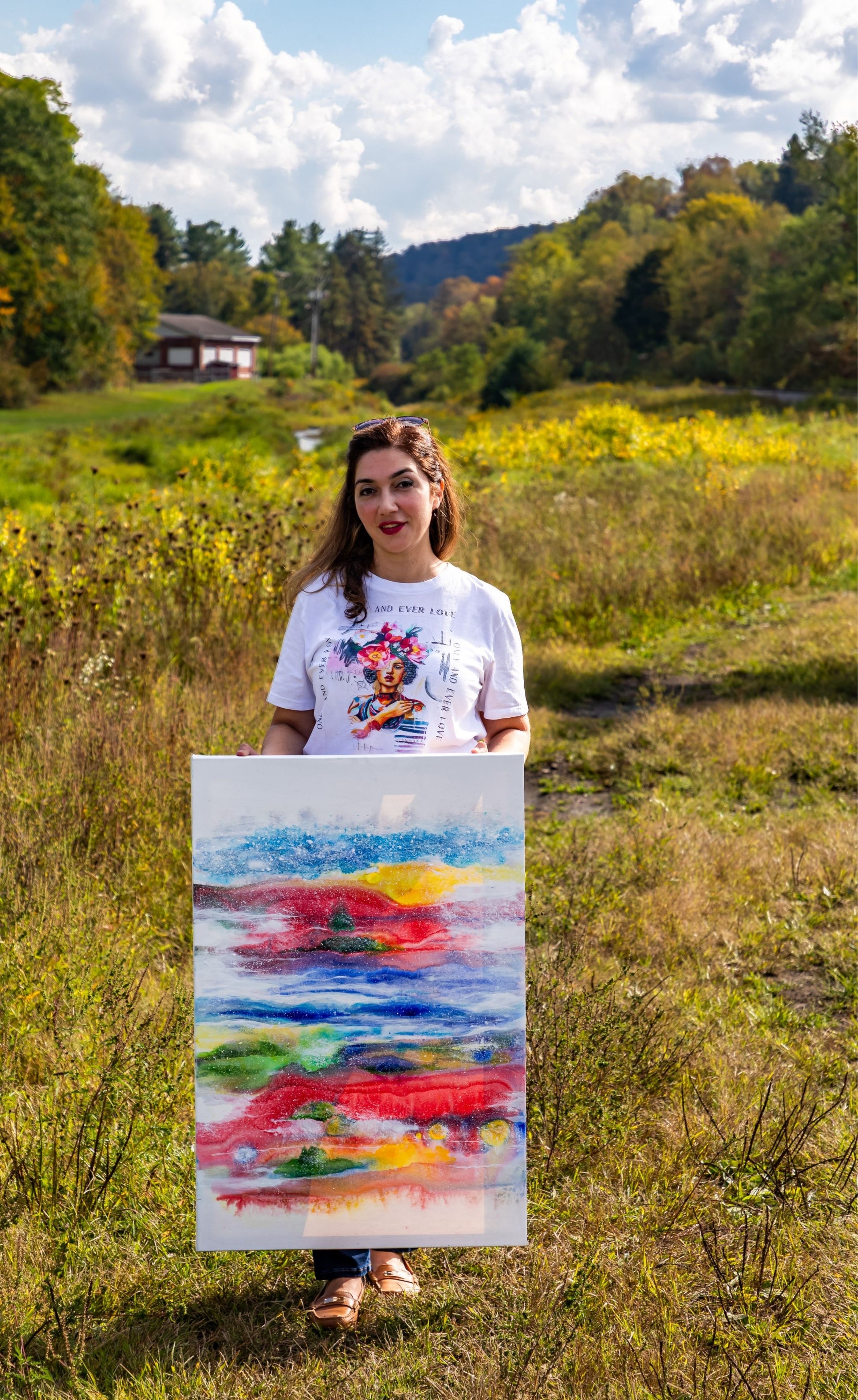 Woman holding a colorful abstract painting in a field with trees and sky in the background